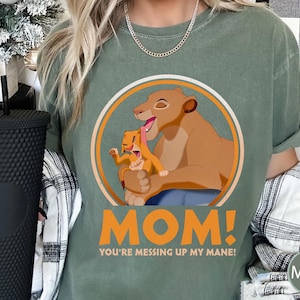 May include: Green t-shirt with a graphic of a lioness and a lion cub. The graphic is in a circle with the text "MOM! You're messing up my mane!" printed below.