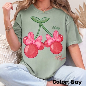 Op de afbeelding: Saliegroen T-shirt met een Minnie Mouse kers ontwerp. De afbeelding toont twee kersen met roze strikken, groene stelen en het Disney-logo. De tekst "Minnie Mouse" staat eronder in cursief.