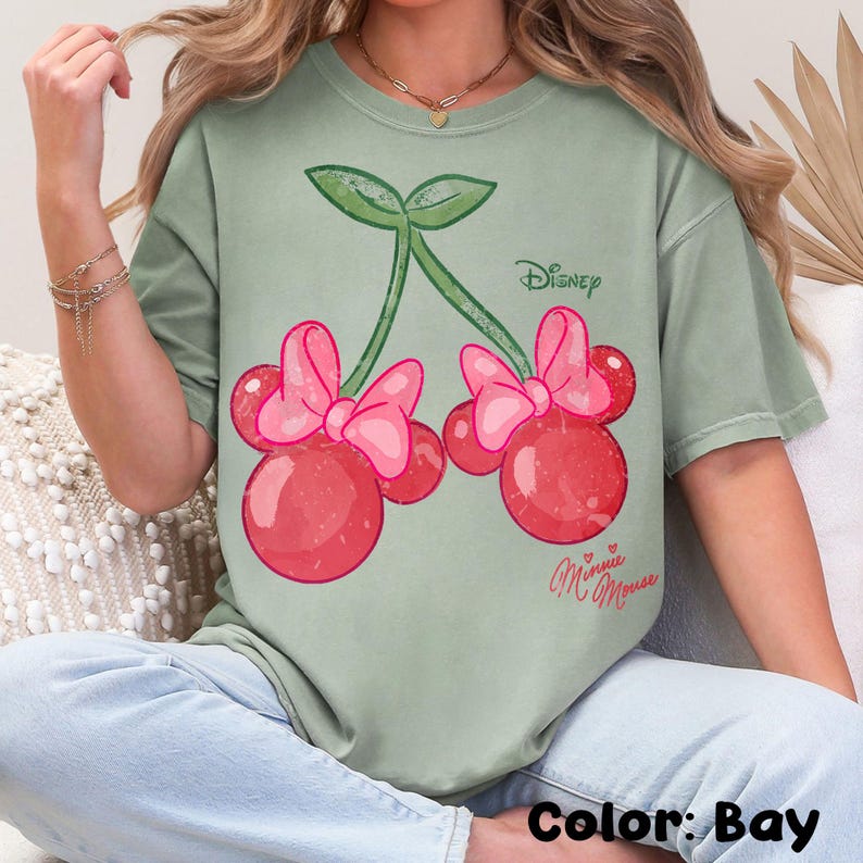 Op de afbeelding: Saliegroen T-shirt met een Minnie Mouse kers ontwerp. De afbeelding toont twee kersen met roze strikken, groene stelen en het Disney-logo. De tekst "Minnie Mouse" staat eronder in cursief.