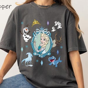 Puede incluir: Una camiseta gris con un estampado gr&aacute;fico de personajes de la pel&iacute;cula Frozen. El gr&aacute;fico presenta a Elsa, Olaf, Sven y una salamandra azul. El texto "Pepper" est&aacute; impreso en la esquina superior izquierda de la camiseta.