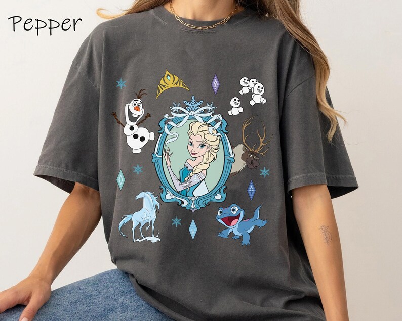 Puede incluir: Una camiseta gris con un estampado gr&aacute;fico de personajes de la pel&iacute;cula Frozen. El gr&aacute;fico presenta a Elsa, Olaf, Sven y una salamandra azul. El texto "Pepper" est&aacute; impreso en la esquina superior izquierda de la camiseta.