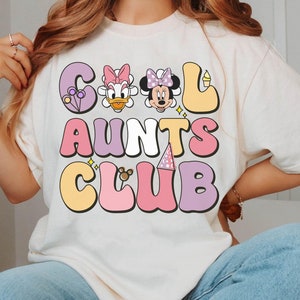Peut inclure: T-shirt blanc avec un motif graphique qui dit "Cool Aunts Club" en lettres colorées en forme de bulles. Le graphique comprend également des images de dessins animés de Minnie Mouse et Daisy Duck.
