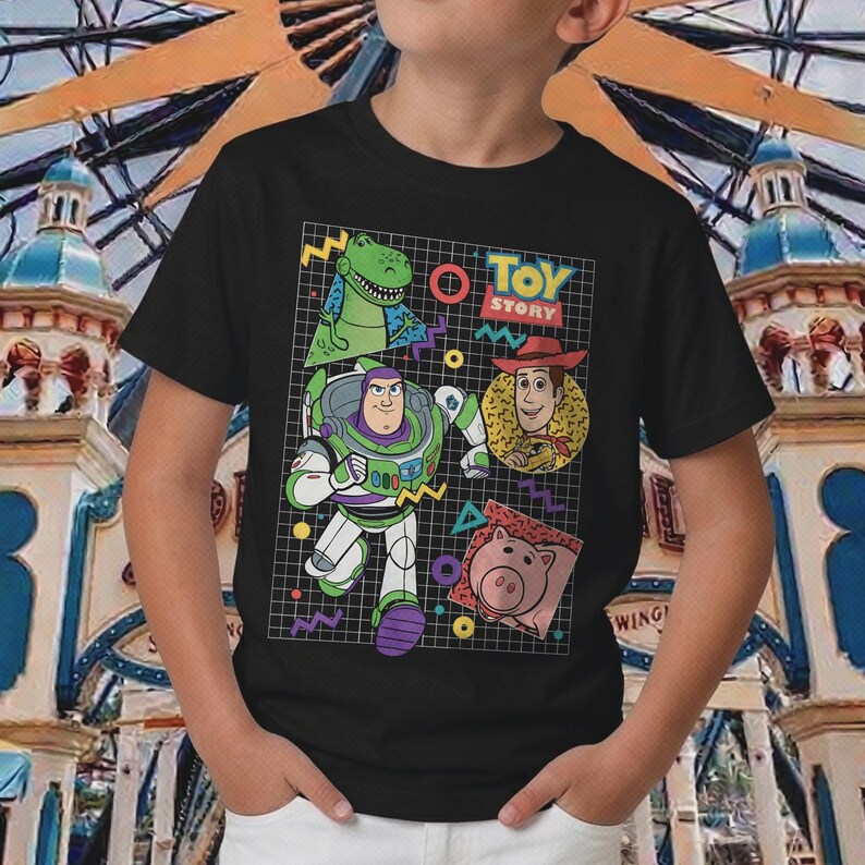 May include: Black t-shirt featuring a colorful graphic of Toy Story characters, including Buzz Lightyear, Woody, Rex, and Hamm. The design includes the words "Toy Story" in a retro style, with geometric shapes and a grid background.
