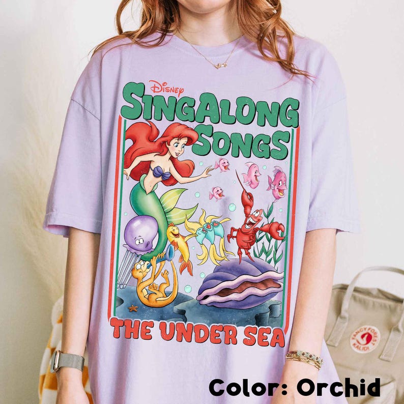 Pu&ograve; includere: T-shirt color orchidea con una grafica colorata della serie "Sing Along Songs" di Disney. Il design include Ariel la sirena, un granchio rosso e altre creature marine. Il testo "Sing Along Songs" e "The Under Sea" &egrave; anch'esso presente.