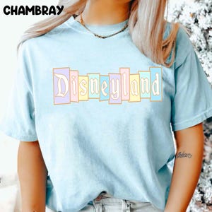 Puede incluir: Camiseta azul claro con la palabra "Disneyland" en colores pastel. El texto est&aacute; en un dise&ntilde;o de bloques verticales. La camiseta tiene mangas cortas y cuello redondo. La palabra "Chambray" est&aacute; impresa en la parte superior izquierda.