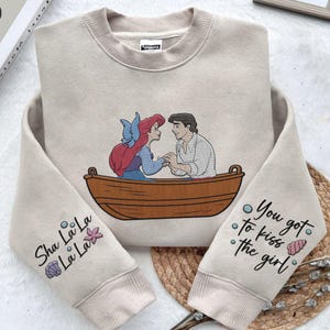May include: A beige sweatshirt featuring a printed design of Ariel and Prince Eric from The Little Mermaid in a boat. The text "Sha La La La La" is printed on one sleeve and "You got to kiss the girl" is printed on the other sleeve.