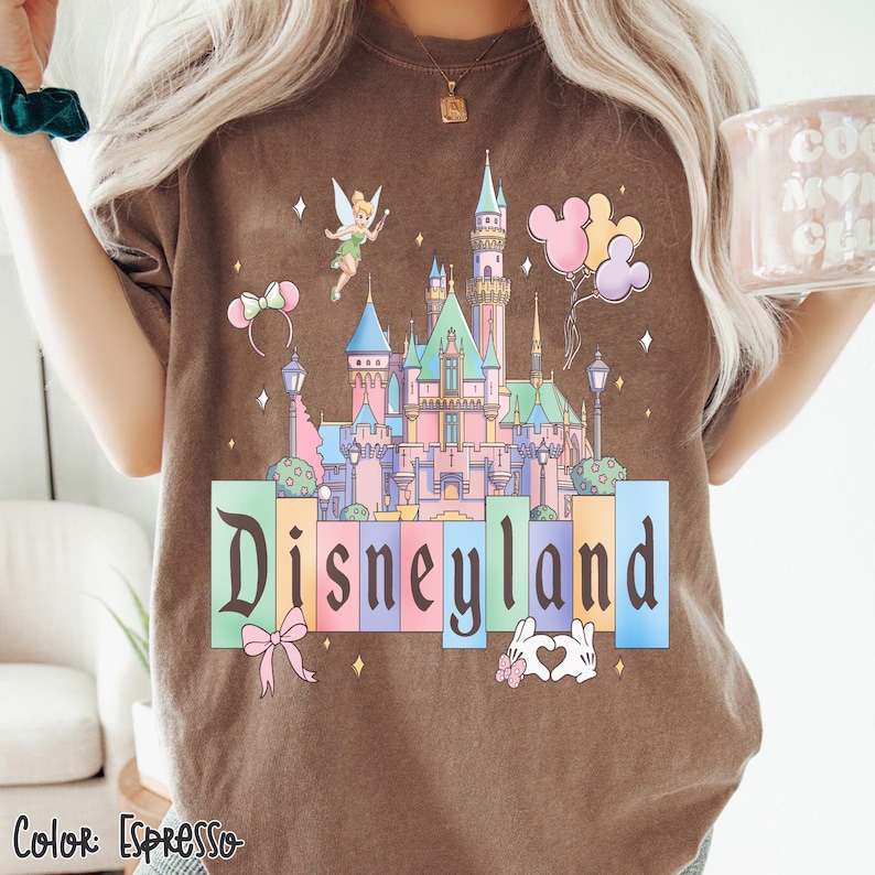 Puede incluir: Camiseta color espresso con un gr&aacute;fico colorido del castillo de Disneylandia con Campanilla, globos y la palabra "Disneyland". El dise&ntilde;o incluye colores pastel y un lazo. La camiseta es de estilo casual y c&oacute;modo.