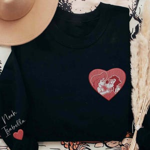 Puede incluir: Sudadera negra con un parche de corazón rojo con una pareja de dibujos animados. La manga izquierda tiene los nombres "Noah Isabella" bordados en cursiva, con un pequeño corazón rojo debajo. Se ve un sombrero color canela.