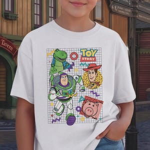 May include: White t-shirt featuring a colorful graphic of characters from the animated film "Toy Story." The design includes Buzz Lightyear, Woody, Rex, and Hamm, set against a grid background with the words "Toy Story."
