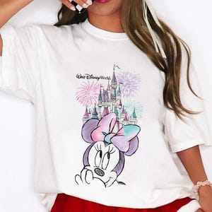Pode incluir: T-shirt branco com um desenho da Minnie Mouse, um castelo e fogos de artif&iacute;cio. O texto "Walt Disney World" est&aacute; acima do castelo. O design &eacute; em tons de roxo, rosa e azul.