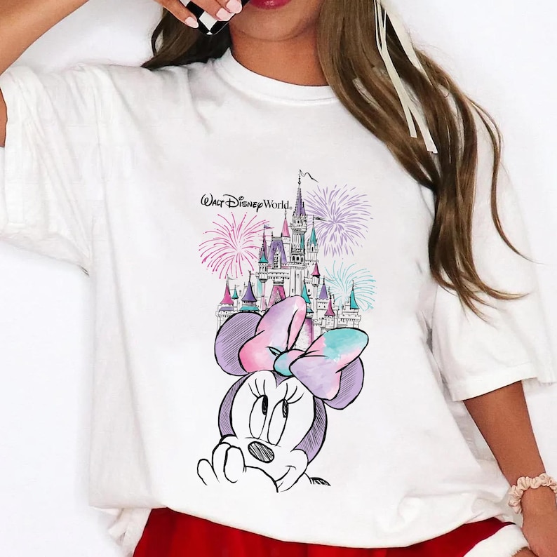 Pode incluir: T-shirt branco com um desenho da Minnie Mouse, um castelo e fogos de artif&iacute;cio. O texto "Walt Disney World" est&aacute; acima do castelo. O design &eacute; em tons de roxo, rosa e azul.