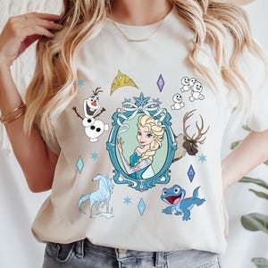 Puede incluir: Una camiseta blanca con un estampado gr&aacute;fico azul y blanco de personajes de la pel&iacute;cula Frozen. El gr&aacute;fico incluye a Elsa, Olaf, Sven y una salamandra azul. El gr&aacute;fico tambi&eacute;n incluye una corona, copos de nieve y un marco.