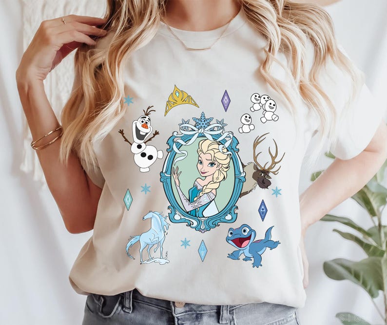 Puede incluir: Una camiseta blanca con un estampado gr&aacute;fico azul y blanco de personajes de la pel&iacute;cula Frozen. El gr&aacute;fico incluye a Elsa, Olaf, Sven y una salamandra azul. El gr&aacute;fico tambi&eacute;n incluye una corona, copos de nieve y un marco.