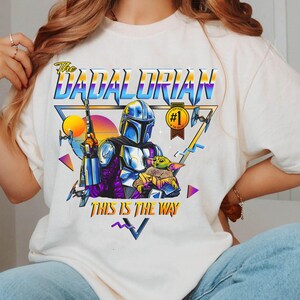May include: White t-shirt with a retro-style graphic of a Mandalorian character holding a baby Yoda. The graphic includes the text "The Dadalorian" and "This is the Way".