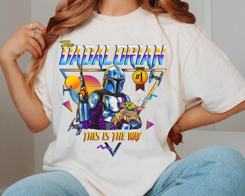 May include: White t-shirt with a retro-style graphic of a Mandalorian character holding a baby Yoda. The graphic includes the text "The Dadalorian" and "This is the Way".