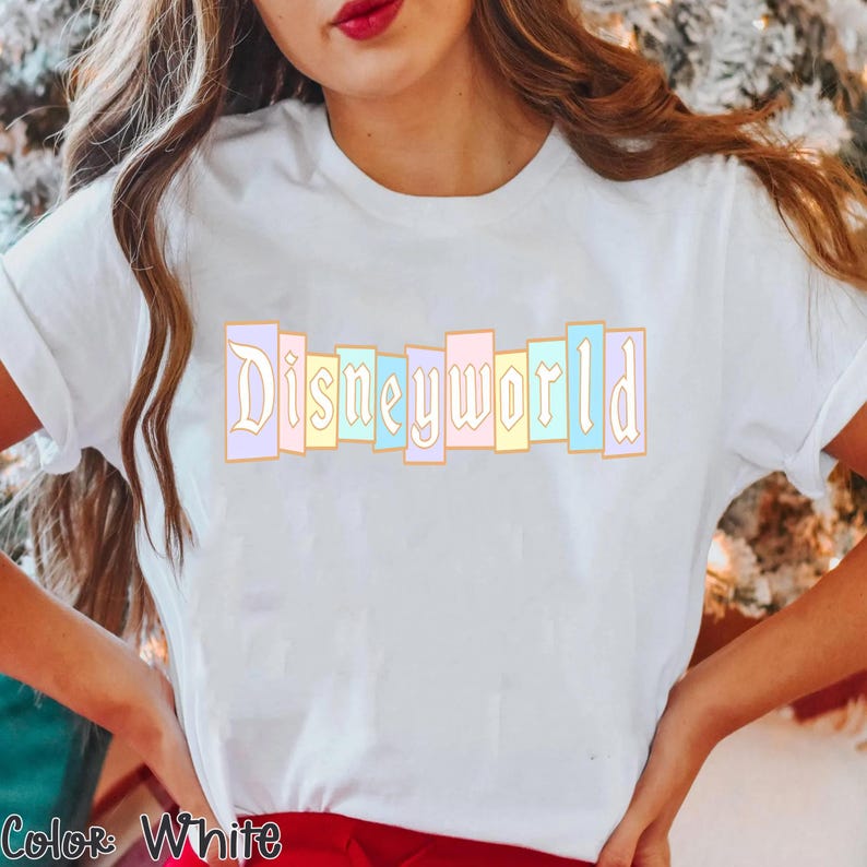 Puede incluir: Camiseta blanca con la palabra "Disneyworld" en colores pastel. El texto est&aacute; en una fuente decorativa y est&aacute; dispuesto verticalmente. La camiseta tiene mangas cortas.