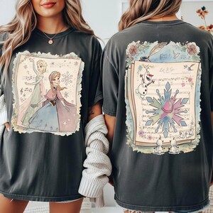May include: Dark gray t-shirts featuring a colorful illustration from the movie Frozen. The front shows Elsa and Anna, while the back displays Olaf, a snowflake, and sheet music with the words "Let It Go."