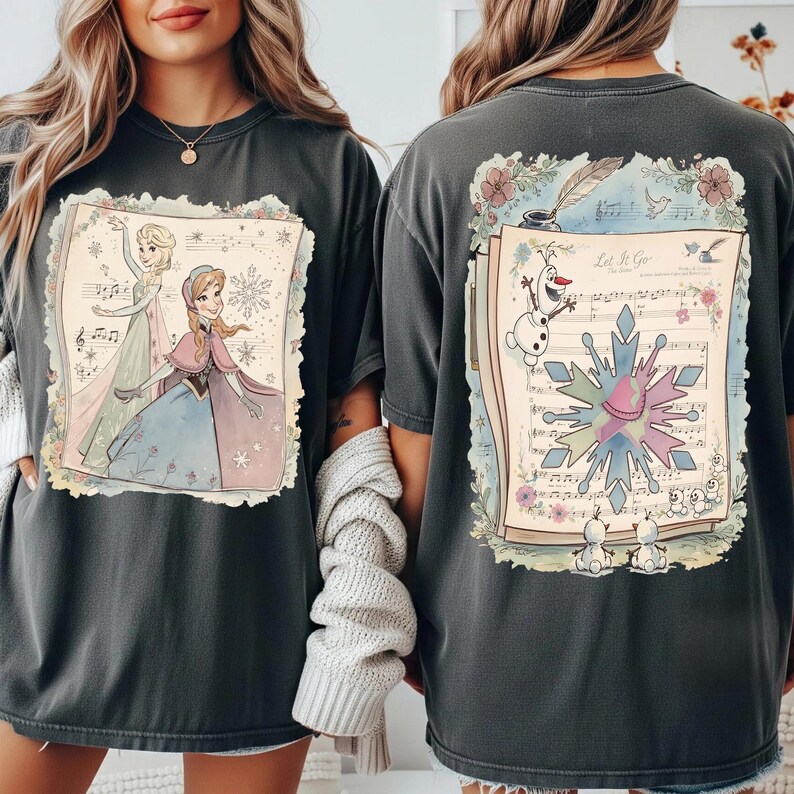 May include: Dark gray t-shirts featuring a colorful illustration from the movie Frozen. The front shows Elsa and Anna, while the back displays Olaf, a snowflake, and sheet music with the words "Let It Go."