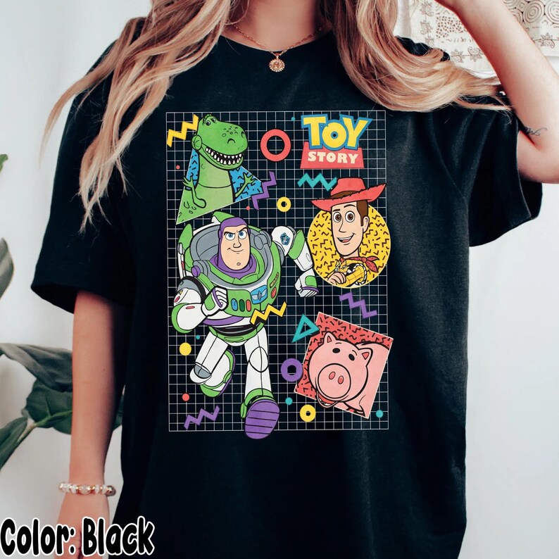 May include: Black t-shirt featuring a colorful graphic of Toy Story characters, including Buzz Lightyear, Woody, Rex, and Hamm. The design is set against a grid background with geometric shapes and the words "Toy Story".