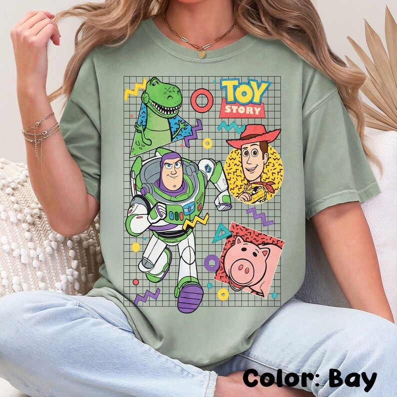 May include: A light green t-shirt featuring a graphic of characters from the animated film "Toy Story." The design includes Buzz Lightyear, Woody, Rex, and Hamm, set against a grid background with colorful geometric shapes. The text "TOY STORY" is also visible.