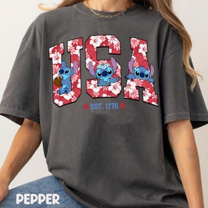 May include: Dark gray t-shirt featuring the word "USA" in red and white floral print, with three blue cartoon characters inside the letters. The text "EST. 1776" is below the letters. The shirt is labeled "PEPPER" at the bottom.