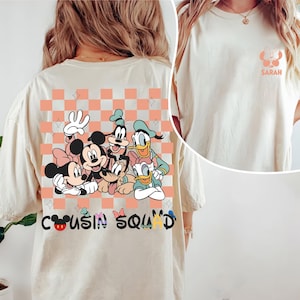May include: A cream-coloured t-shirt with a graphic of Disney characters, including Mickey and Minnie Mouse, Goofy, and Donald Duck, with the text "Cousin Squad." A smaller graphic with the name "Sarah" is on the front.