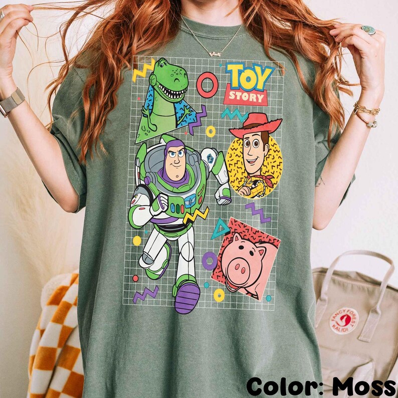 May include: Moss green t-shirt featuring a colorful graphic of Toy Story characters, including Rex, Buzz Lightyear, Woody, and Hamm. The design is set against a grid pattern with geometric shapes and the words "Toy Story".