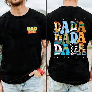 Op de afbeelding: Zwart T-shirt met de tekst "DAD LIFE" in een kleurrijk ontwerp op de voorkant. De achterkant van het shirt heeft het woord "DADA" in een speelse, veelkleurige lettertype met steraccenten, wat een leuk en opvallend ontwerp cre&euml;ert.
