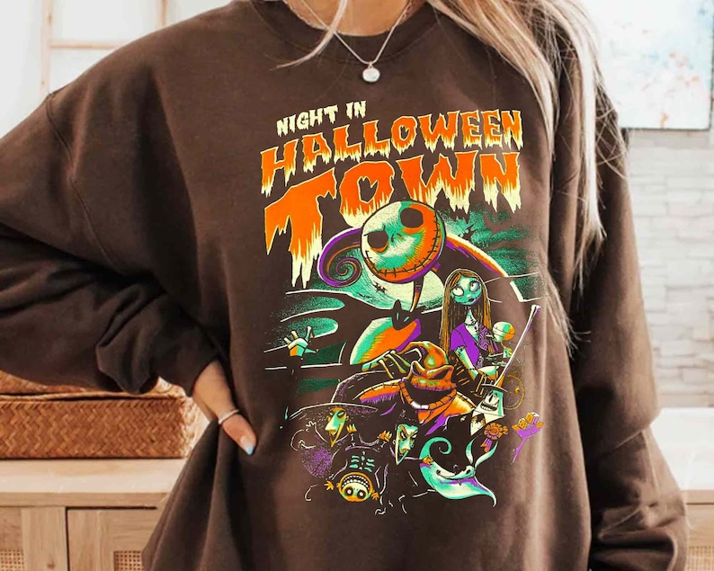 Może przedstawiać: Brązowa bluza z nadrukiem graficznym postaci z filmu "Koszmar przed świętami". Na bluzie nadrukowany jest tekst "Night in Halloween Town".
