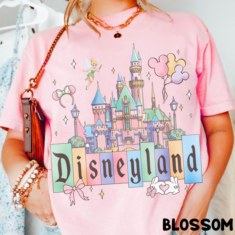 Puede incluir: Camiseta rosa con un dise&ntilde;o del castillo de Disneyland, Campanilla, globos de Mickey Mouse y la palabra "Disneyland". La camiseta tiene cuello redondo y mangas cortas.