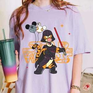 May include: Purple t-shirt with a cartoon image of a tiger wearing a Darth Vader costume and holding a lightsaber. The tiger is holding a Mickey Mouse head and two Death Star ornaments. The text "Star Wars" is printed on the shirt.