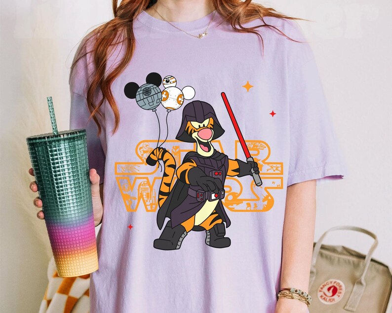 May include: Purple t-shirt with a cartoon image of a tiger wearing a Darth Vader costume and holding a lightsaber. The tiger is holding a Mickey Mouse head and two Death Star ornaments. The text "Star Wars" is printed on the shirt.