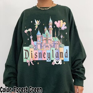 Puede incluir: Sudadera verde bosque con un gr&aacute;fico de Disneyland. El dise&ntilde;o incluye un castillo, Campanilla, globos y la palabra "Disneyland" en letras de colores. Se ve un collar de cadena dorada.