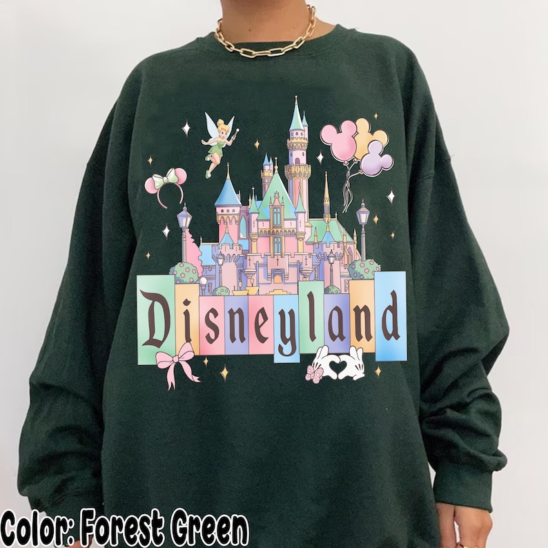 Puede incluir: Sudadera verde bosque con un gr&aacute;fico de Disneyland. El dise&ntilde;o incluye un castillo, Campanilla, globos y la palabra "Disneyland" en letras de colores. Se ve un collar de cadena dorada.