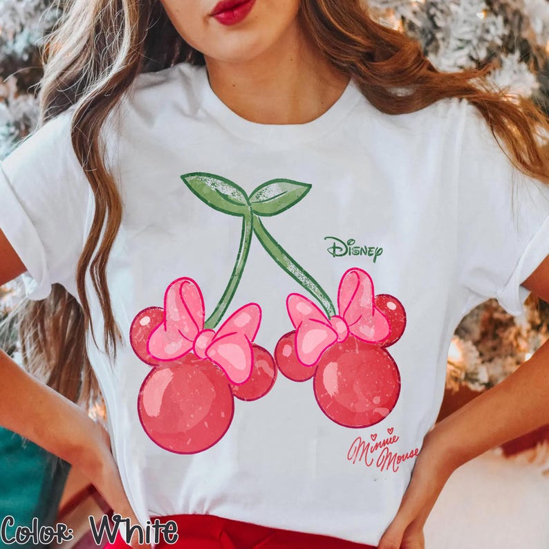 Op de afbeelding: Wit T-shirt met een speels ontwerp van kersen versierd met Minnie Mouse strikken. De kersen zijn rood met roze strikken en groene stelen. Het Disney-logo staat boven de kersen gedrukt en de handtekening van Minnie Mouse staat eronder.