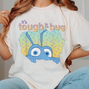 以下が含まれることがあります： 「It's tough to be a bug」というテキストと、アニメーションの蟻のグラフィックが描かれた白いTシャツです。