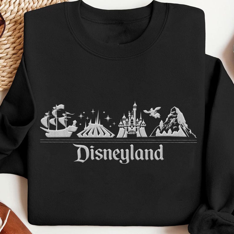 Op de afbeelding: Zwart sweatshirt met een wit geborduurd Disneyland-ontwerp. Het ontwerp omvat een schip, de Matterhorn, een kasteel en het woord "Disneyland". De sweatshirt heeft een ronde hals.
