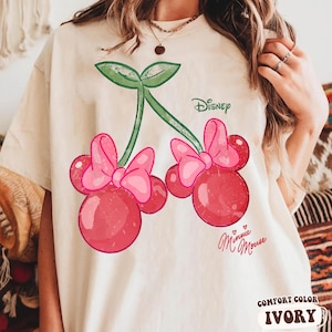 Op de afbeelding: Ivoor kleurig T-shirt met een Minnie Mouse kers ontwerp. De afbeelding toont twee kersen met roze strikken, groene stelen en het Disney-logo. De woorden "Minnie Mouse" staan eronder in cursief. Het shirt is gelabeld "Comfort Color Ivory".