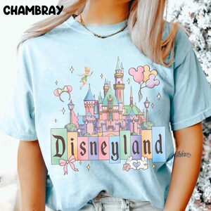 Puede incluir: Camiseta azul claro con una ilustraci&oacute;n colorida de un castillo, Campanilla y globos de Mickey Mouse. La palabra "Disneyland" est&aacute; impresa en arco&iacute;ris. La camiseta tiene mangas cortas y cuello redondo.