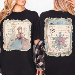 May include: Black t-shirt featuring a colorful illustration of Elsa and Anna from Frozen, with musical notes and a snowflake design. The back of the shirt shows Olaf, a snowflake, and the text "Let It Go".
