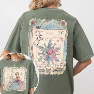 May include: Olive green t-shirt featuring a graphic of a musical score with the words "Let It Go" and a snowflake design. The design includes illustrations of Olaf and other characters from the movie.