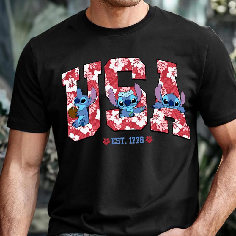 May include: Black t-shirt featuring the word "USA" in a red floral pattern with three cartoon characters. The text "EST. 1776" is below the word "USA". The characters are blue with large eyes.