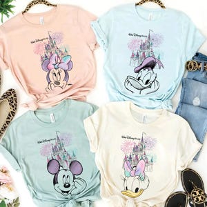Pode incluir: Quatro camisetas em tons pastel com personagens da Disney e o texto "Walt Disney World". Cada camisa exibe um personagem diferente: Minnie Mouse, Pato Donald, Mickey Mouse e Margarida, com um castelo e fogos de artif&iacute;cio.