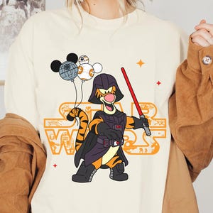 May include: A white t-shirt with a cartoon image of Tigger dressed as Darth Vader. Tigger is holding a lightsaber and has a black cape. There are three small cartoon images of Mickey Mouse, R2-D2, and a Death Star above Tigger's head. The text "Star Wars" is printed in orange and black on the shirt.
