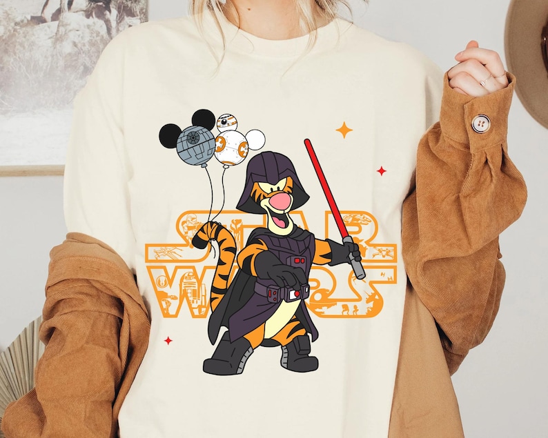 May include: A white t-shirt with a cartoon image of Tigger dressed as Darth Vader. Tigger is holding a lightsaber and has a black cape. There are three small cartoon images of Mickey Mouse, R2-D2, and a Death Star above Tigger's head. The text "Star Wars" is printed in orange and black on the shirt.