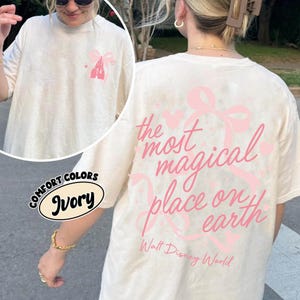 May include: Ivory-colored Comfort Colors t-shirt with a pink castle graphic on the front and the phrase "the most magical place on earth" on the back. The text "Walt Disney World" is also printed on the back.