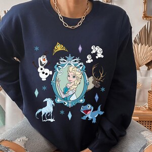 Puede incluir: Una sudadera azul marino con un estampado gr&aacute;fico de Elsa de Frozen. El estampado incluye a Elsa, Olaf, Sven el reno y algunos mu&ntilde;ecos de nieve. La sudadera tiene cuello redondo y mangas largas.