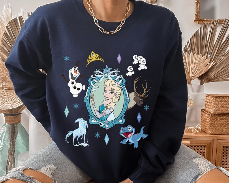 Puede incluir: Una sudadera azul marino con un estampado gr&aacute;fico de Elsa de Frozen. El estampado incluye a Elsa, Olaf, Sven el reno y algunos mu&ntilde;ecos de nieve. La sudadera tiene cuello redondo y mangas largas.