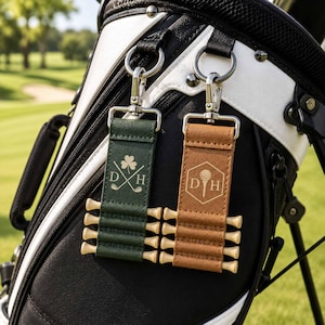 May include: Two rectangular faux leather golf tee holders with silver metal clips. One is dark green with a shamrock and golf club design, the other is tan with a golf ball in a hexagon. Both feature the letters D and H and hold four wooden tees.