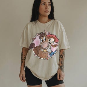 May include: Cream-colored t-shirt featuring a cartoon illustration of a horse hugging a cowgirl. The graphic is framed by a pink circle with stars and a rope border. The cowgirl wears a red hat, blue jeans, and a white shirt.